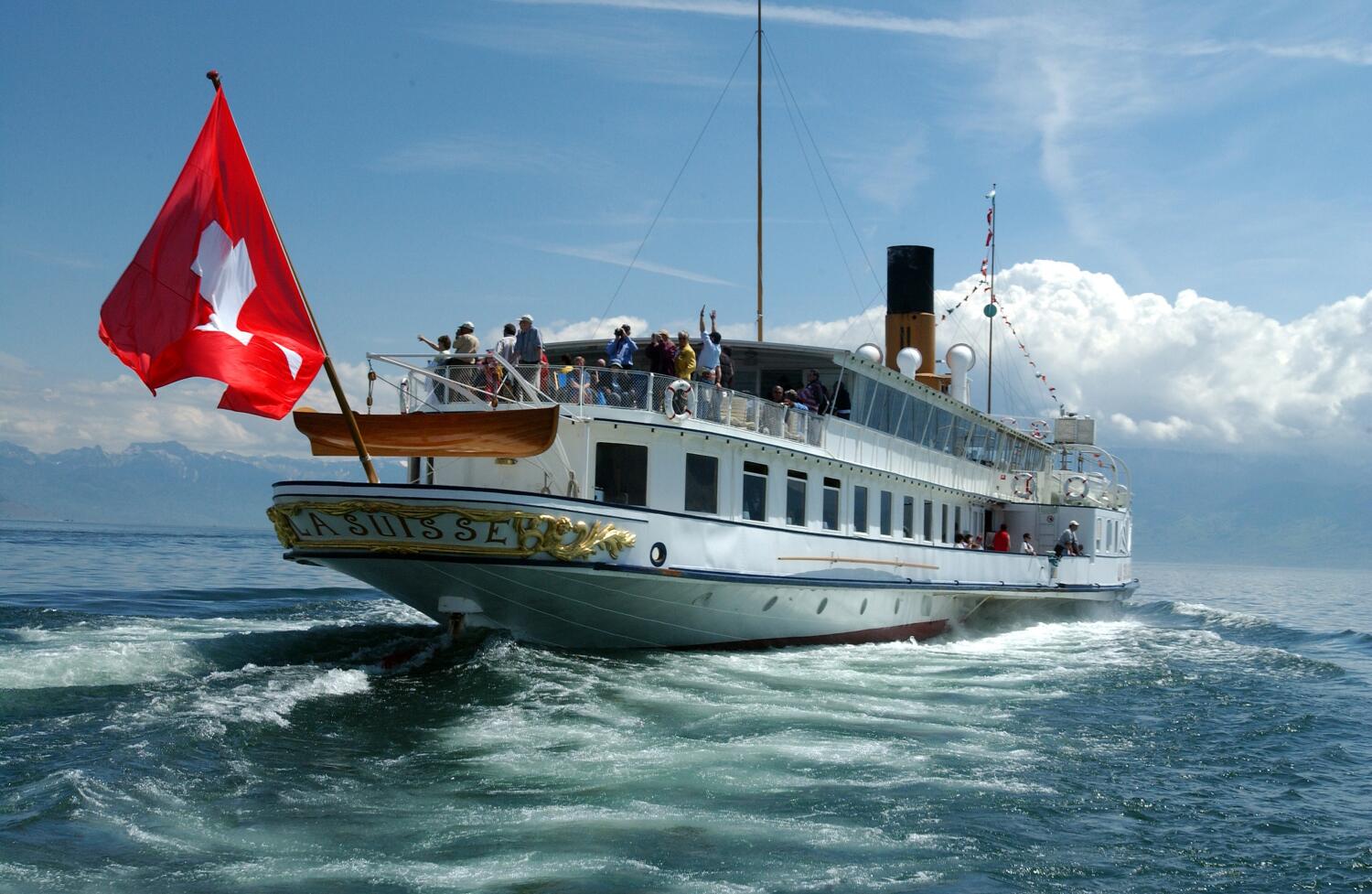 Un bateau CGN blanc navigue sur le lac Léman, vu de l'arrière avec des passagers sur le pont et le drapeau suisse flottant à la poupe